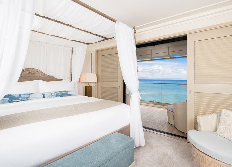 Room with canopy bed, lamp, seating, and balcony with view of ocean.