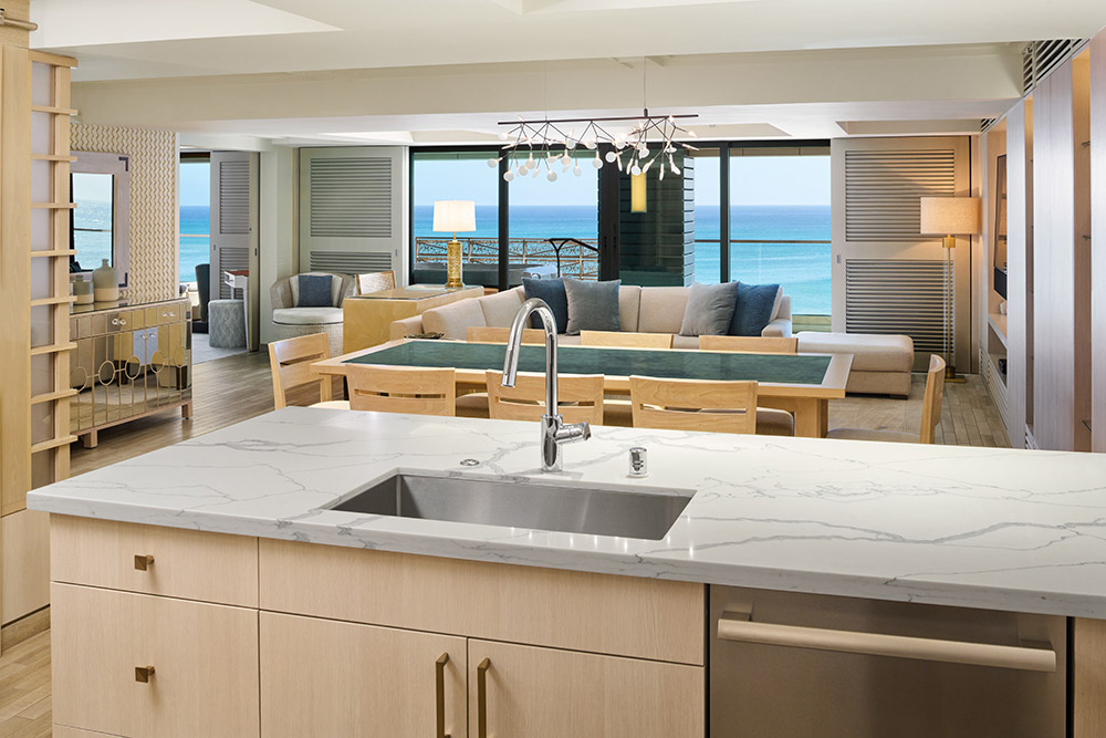 Kitchen sink and countertop seating opens to dining area with table and chairs, and living area with couch and balcony with ocean view.