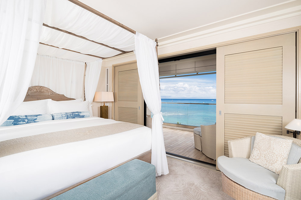 Room with canopy bed, lounge chair, lamp, balcony with ocean view.