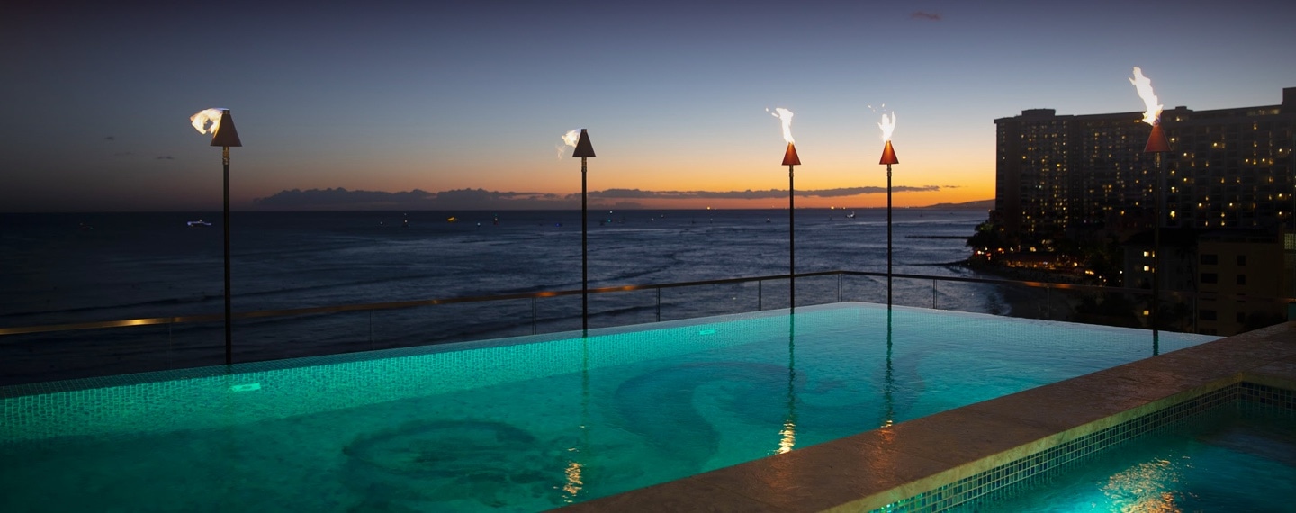 Infinity pool with lit torch lighting, ocean view in background at sunset.