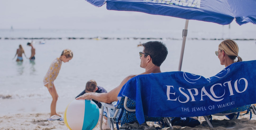 Family relaxing on Waikiki Beach with chairs, umbrella, beach ball, and ESPACIO towel.